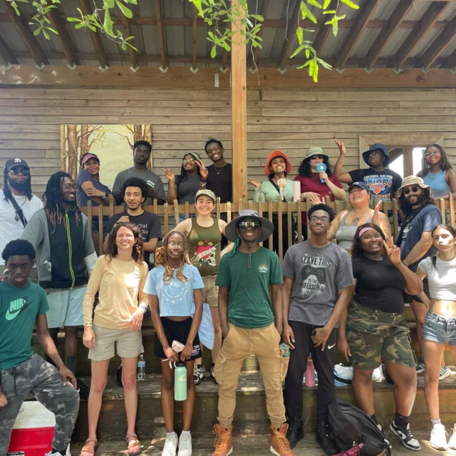 A diverse group of people posing together on wooden steps in front of a rustic building, with trees visible in the background. They appear relaxed and are casually dressed.