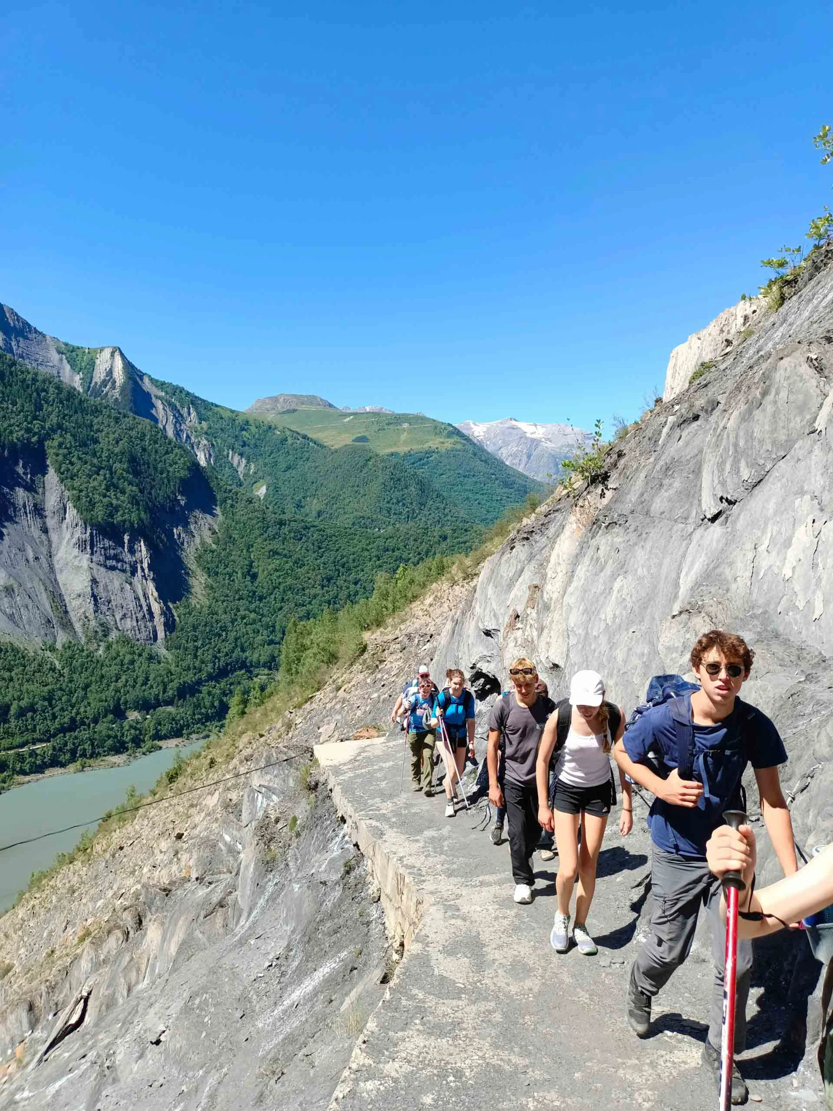 A group of hikers walks along a narrow mountain path with a river and green mountains in the background under a clear blue sky.