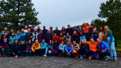 A large group of people pose for a photo outside, standing and kneeling on a dirt path with trees in the background.