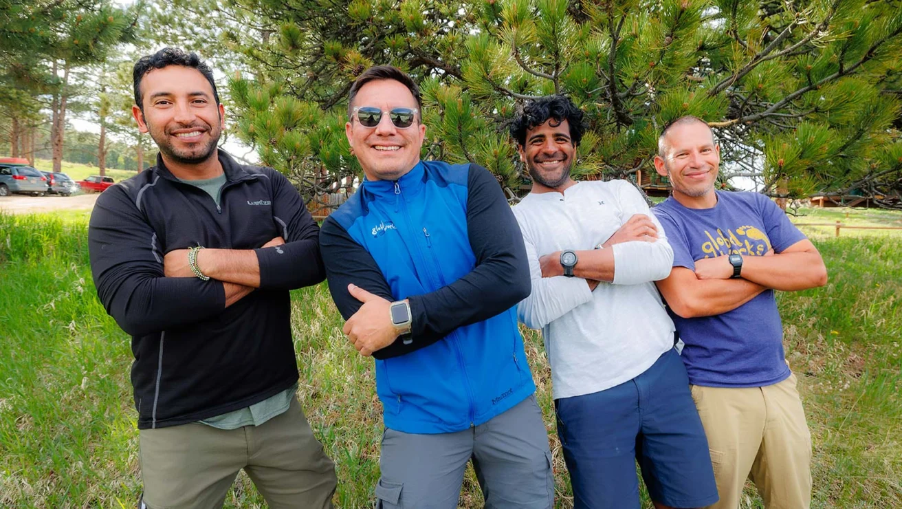 Four men stand outdoors in casual clothing, smiling with arms crossed. They are in front of a tree, surrounded by grass and other trees.