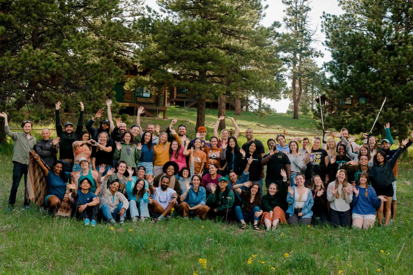A group of people poses together outdoors on a grassy field with trees and a cabin in the background.