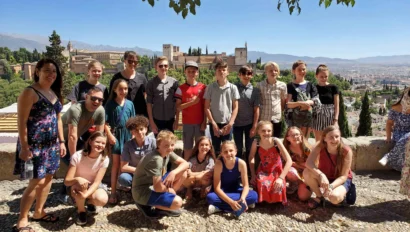 A group of people, including teenagers and adults, gather outdoors on a sunny day with a scenic city and landscape in the background.