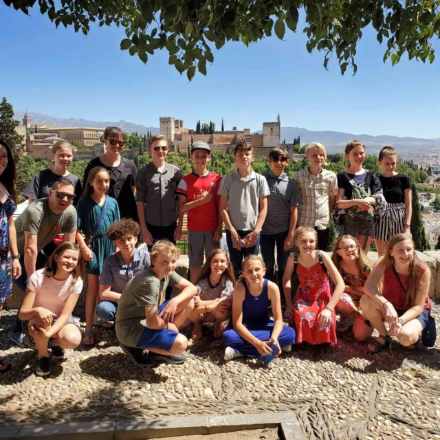 A group of people, including teenagers and adults, gather outdoors on a sunny day with a scenic city and landscape in the background.