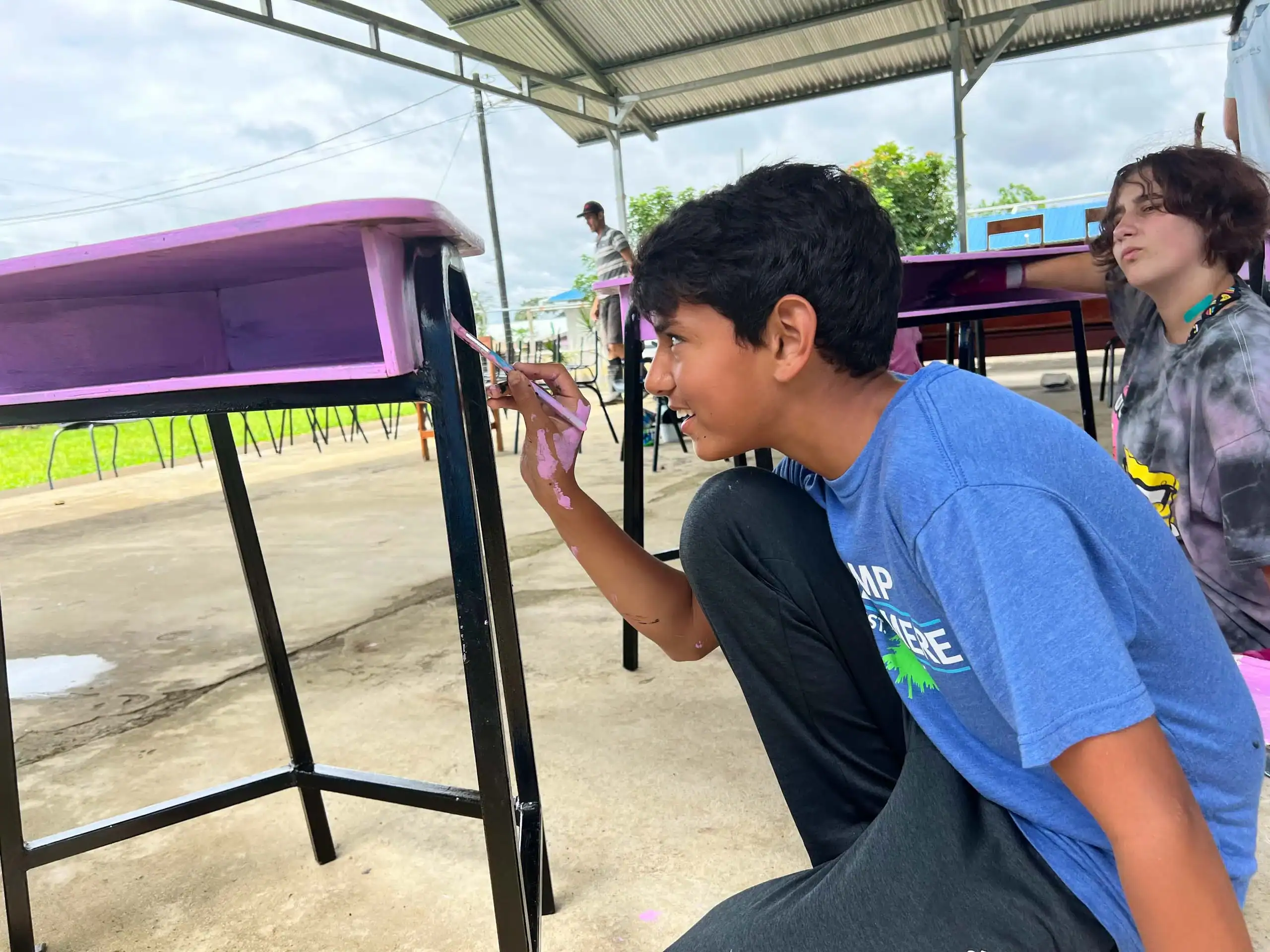 A boy in a blue shirt paints the leg of a pink desk outside under a metal roof during one of the Middle School student trips, while another person sits nearby.