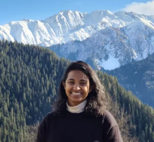 A person smiling in front of snow-covered mountains and pine forests under a clear blue sky.
