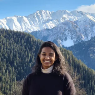 A person smiling in front of snow-covered mountains and pine forests under a clear blue sky.