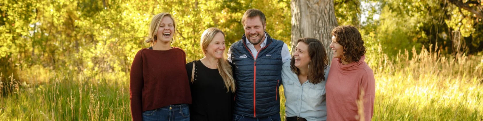 Five people standing outdoors, smiling and laughing in a grassy area with trees and sunlight in the background.