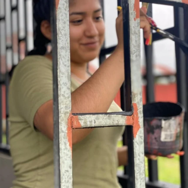 A person paints a metal fence with orange paint using a brush, holding a can of paint in the other hand.
