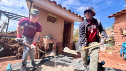 Two people in casual clothing shovel sand on a tarp outside a small building with a tiled roof.
