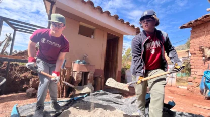 Two people in casual clothing shovel sand on a tarp outside a small building with a tiled roof.