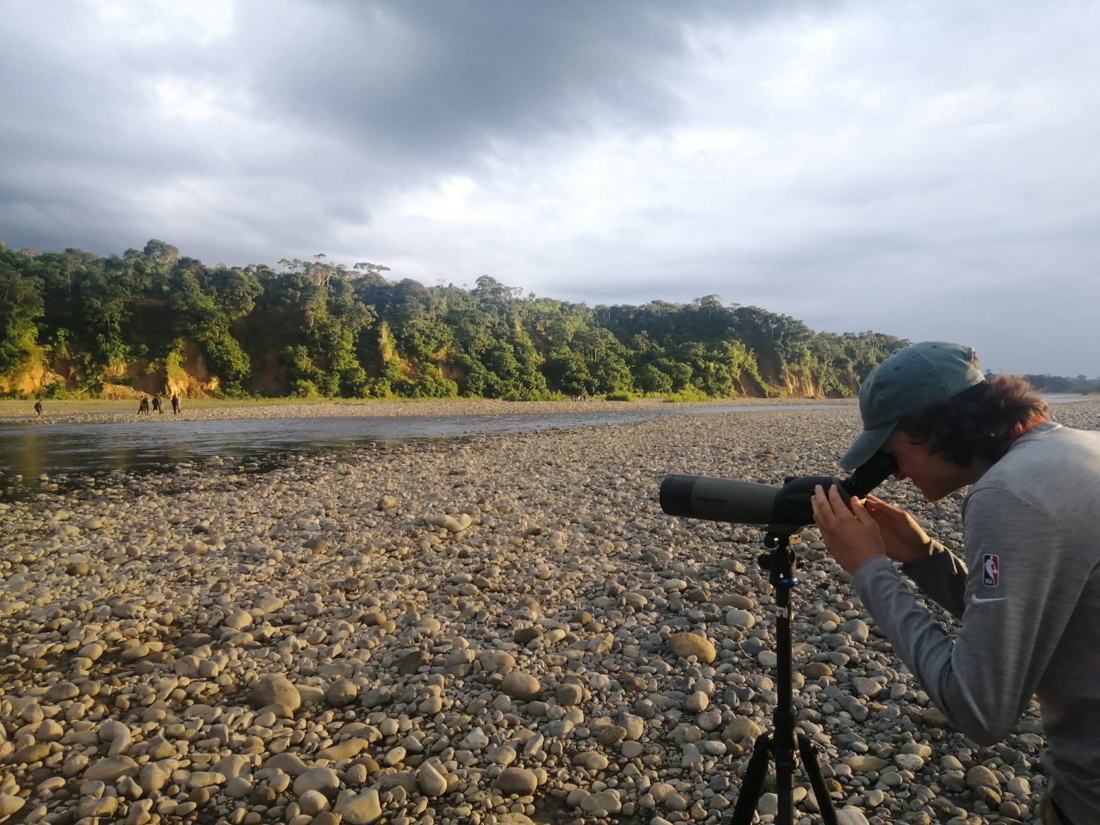 Person using a telescope on a rocky riverbank with a forest in the background under a cloudy sky.