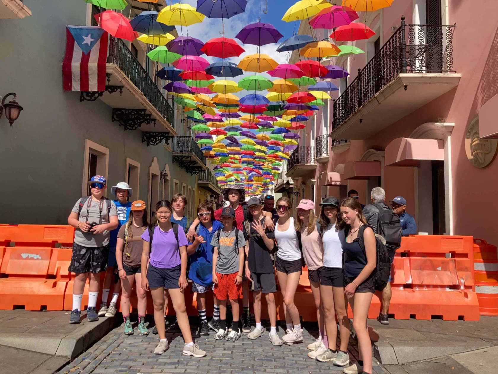 A group of people stand on a cobblestone street under a canopy of colorful umbrellas. Orange barricades line the sides. A Puerto Rican flag is visible on a building.