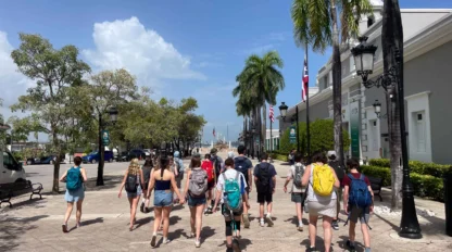 A group of people with backpacks walk down a sunny street lined with palm trees and buildings.