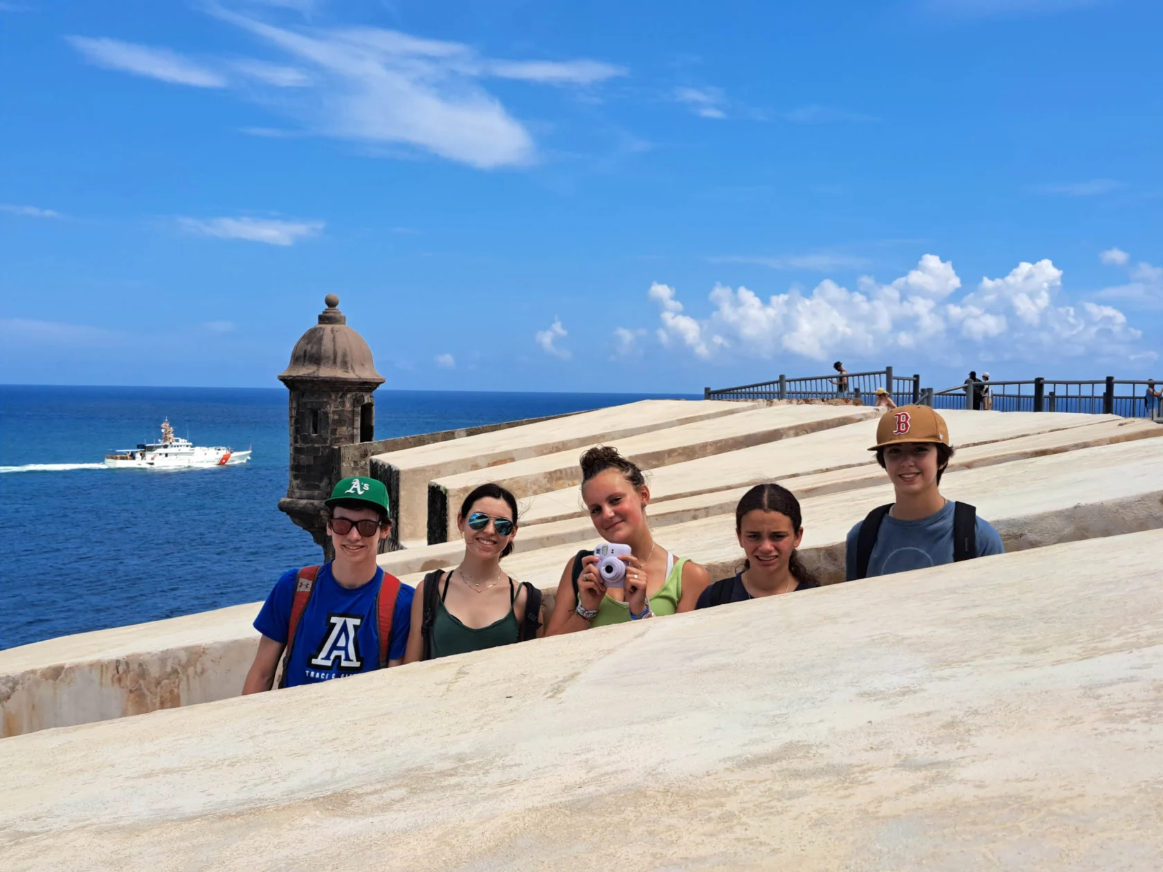 Five people stand on a stone structure near the ocean, with a boat in the background under a blue sky.