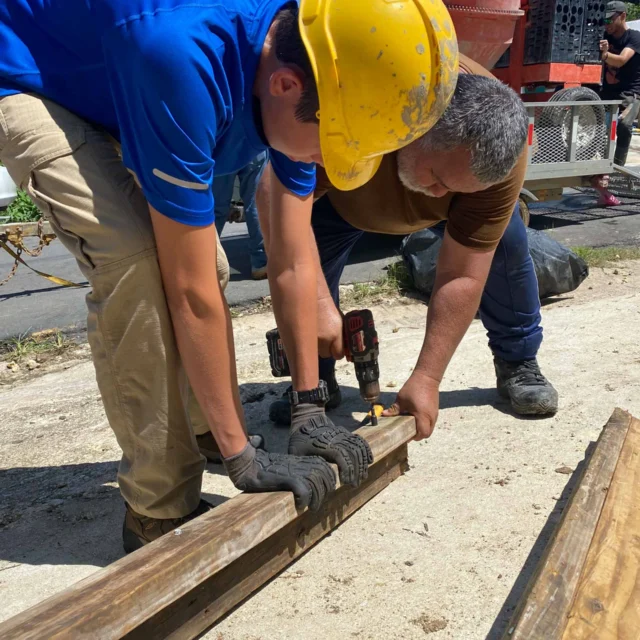 Two people in work gear use power drills to attach wooden beams on a sunny day.