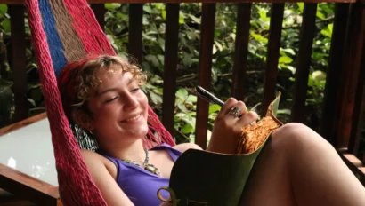 A person lounges in a hammock, smiling and writing in a notebook. They are wearing a purple tank top.