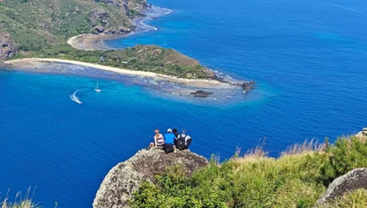 Three people sit on a grassy cliff overlooking a small island with a sandy beach surrounded by clear blue water. Two boats are visible on the water.