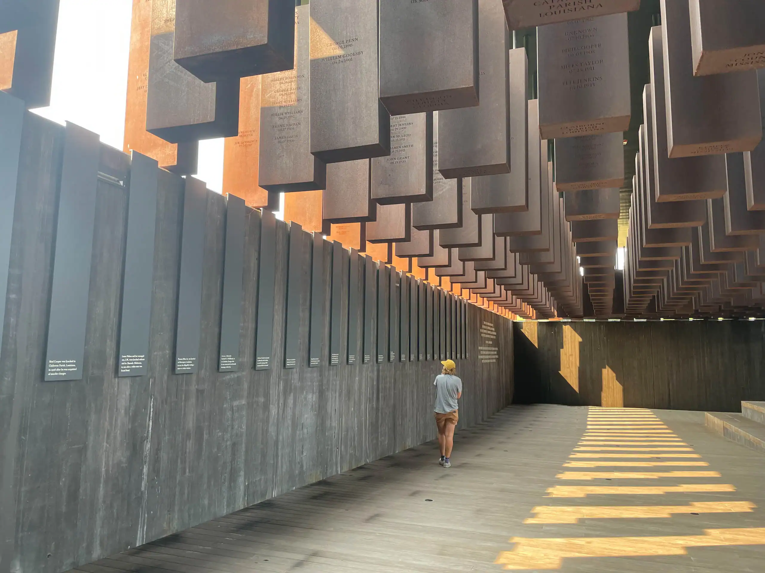 A person walks alone through an outdoor memorial with suspended metal columns and engraved plaques on the walls, exploring a powerful site of American history travel under natural daylight.
