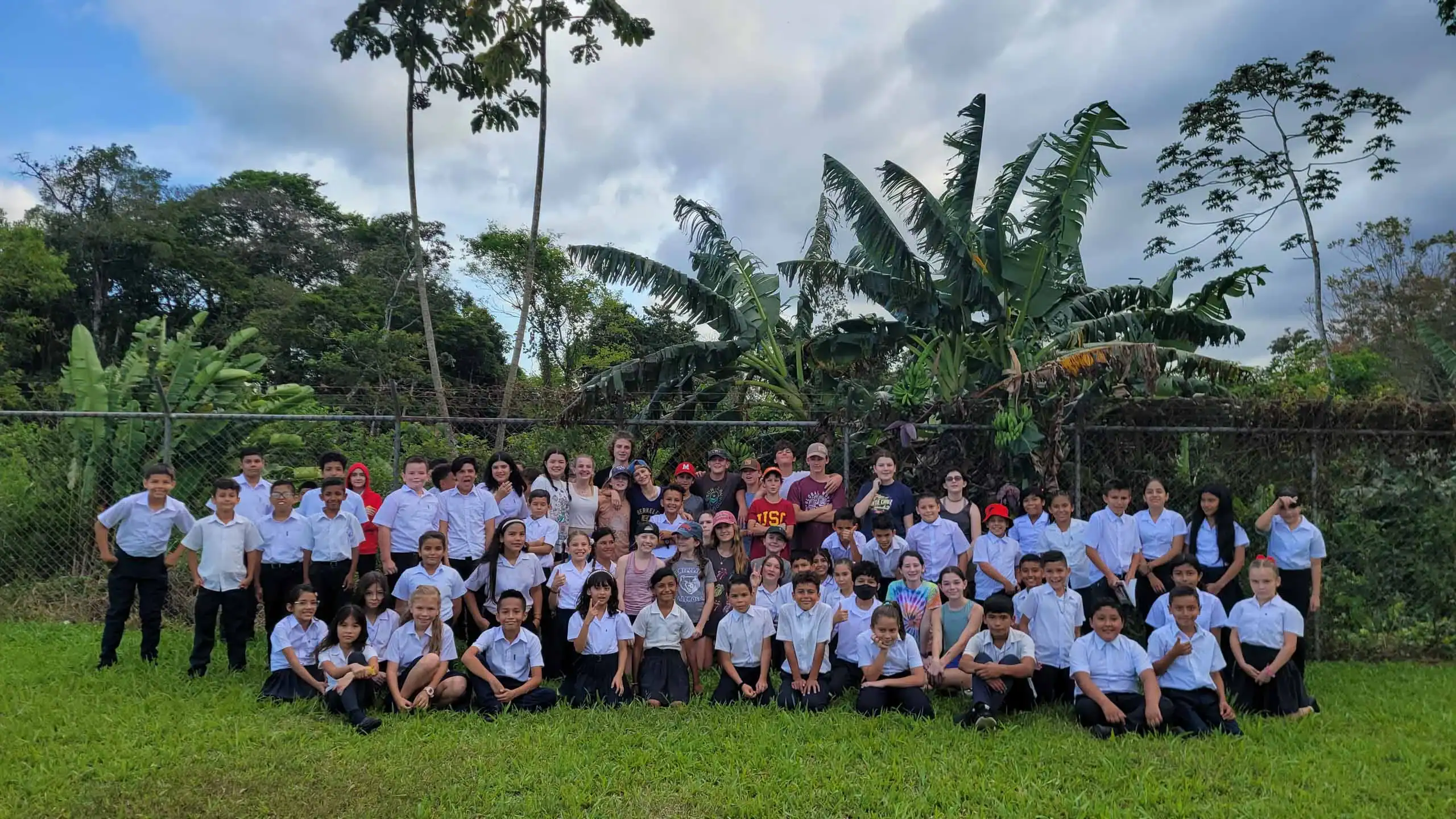 A large group of children and adults pose for a photo outdoors on grass, with tropical trees and plants in the background during their Costa Rica school trip.