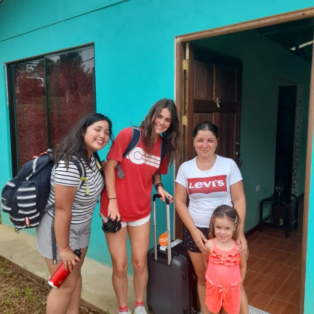 Three women and a girl stand outside a turquoise building. The women have luggage and backpacks, and they are smiling.