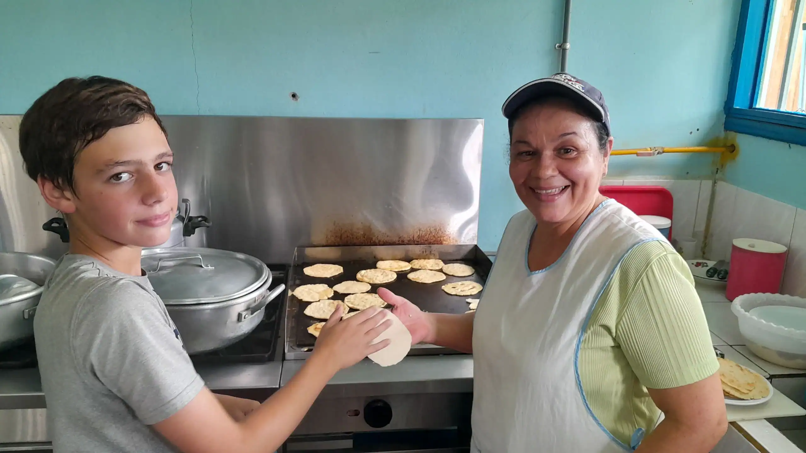 A student traveler and a woman smile while making tortillas on a griddle in a kitchen during their Costa Rica school trip. The woman holds a tortilla, and several cooked tortillas are visible on the stove.