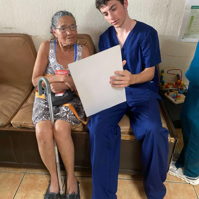 A young student on a medical study abroad school trip in Costa Rica in blue scrubs reviews a document with an older woman in a clinic setting.