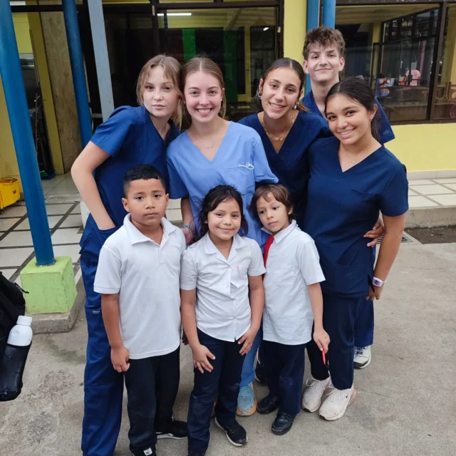 Group of students in scrubs posing with three children in school uniforms outside a building.