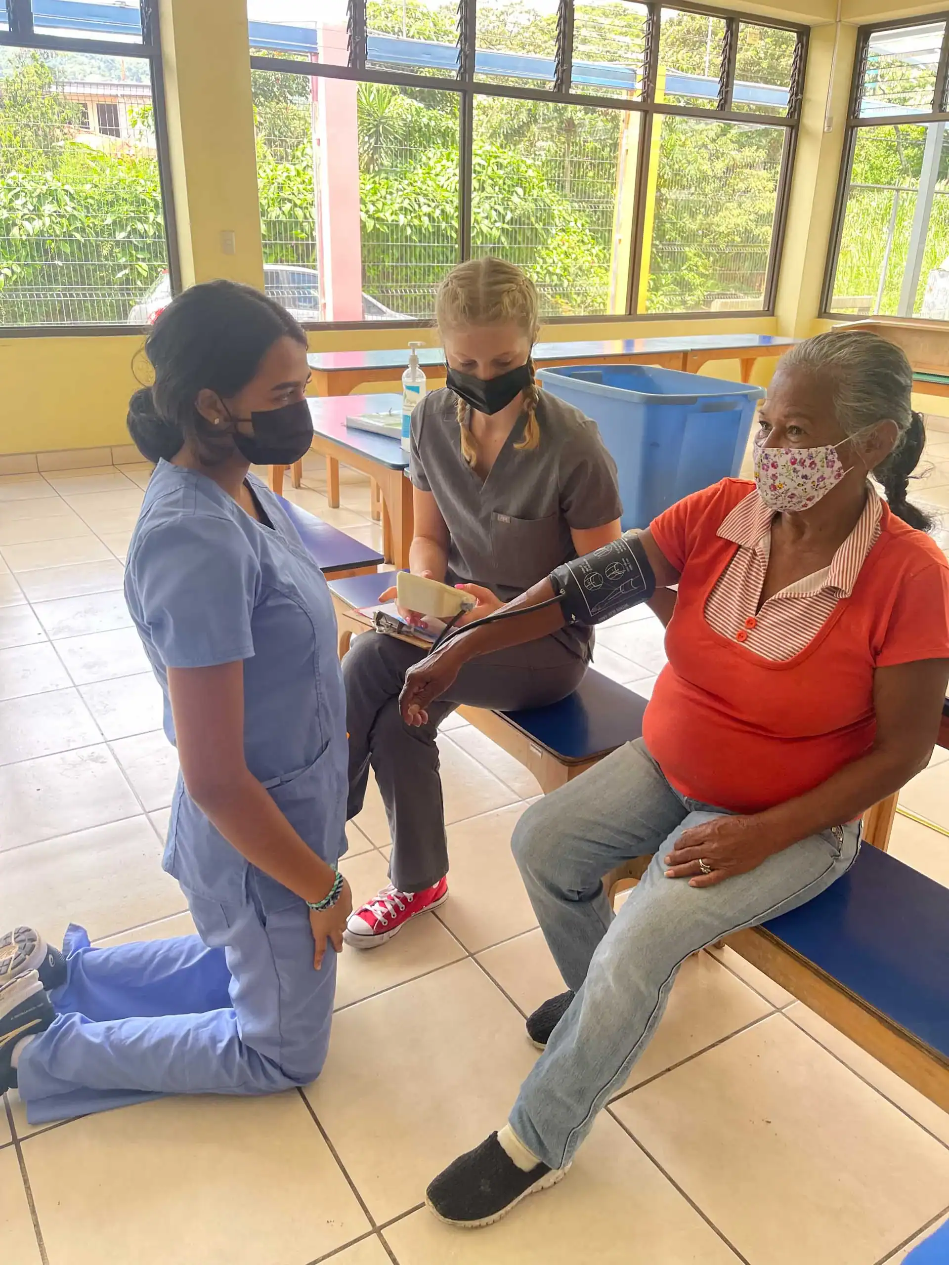 During a teen medical trip abroad, two healthcare workers check the blood pressure of an older woman sitting on a bench in a brightly lit room. All three are wearing face masks.