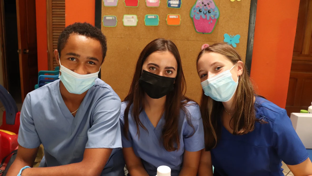 Three people wearing scrubs and masks sit together with colorful objects in the background.