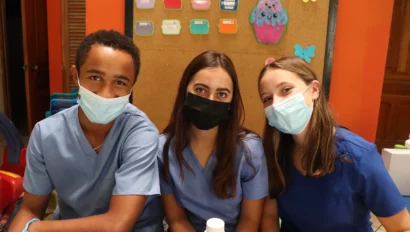 Three people wearing scrubs and masks sit together with colorful objects in the background.