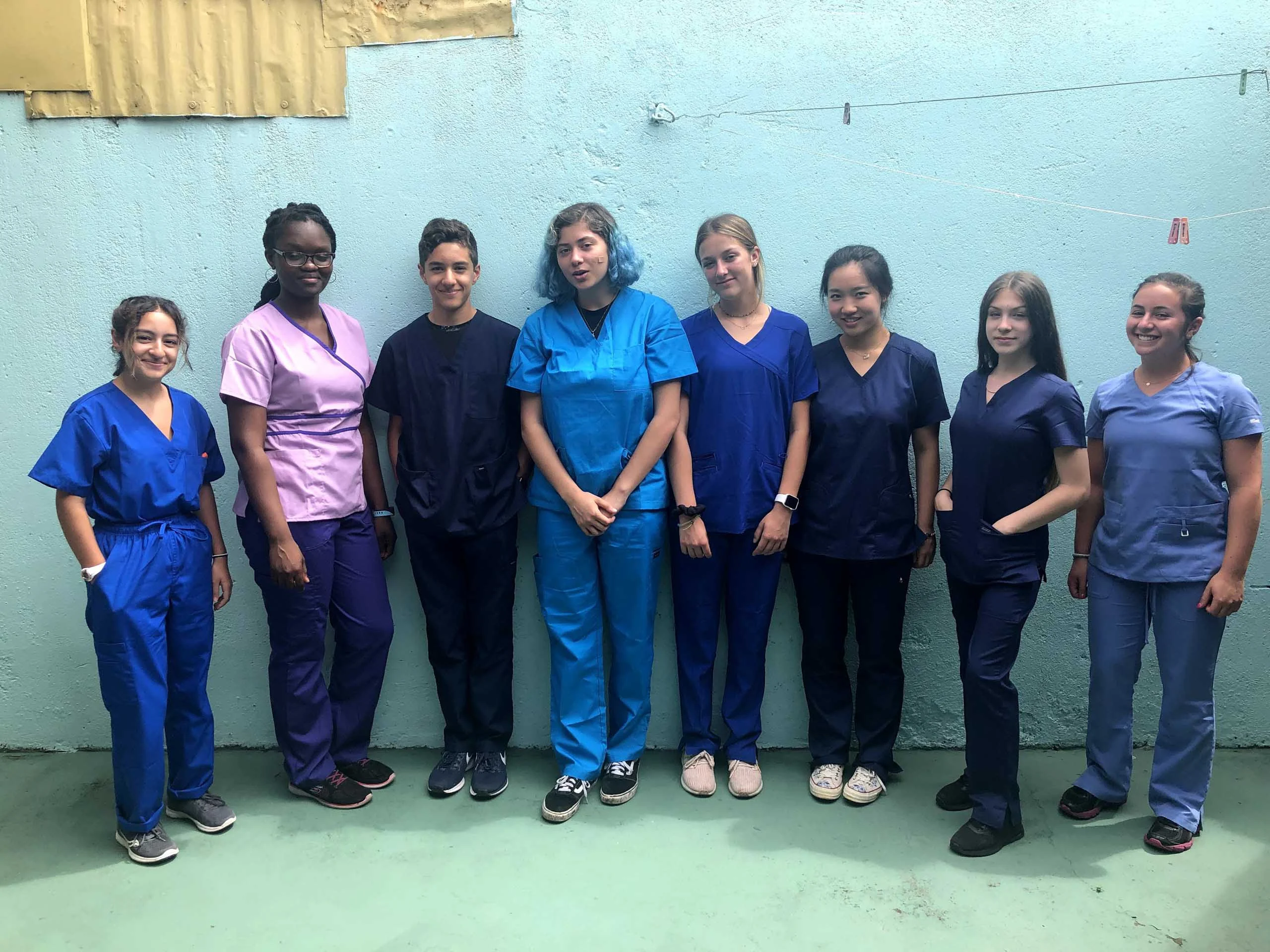 A group of eight people standing in scrubs against a light blue wall.