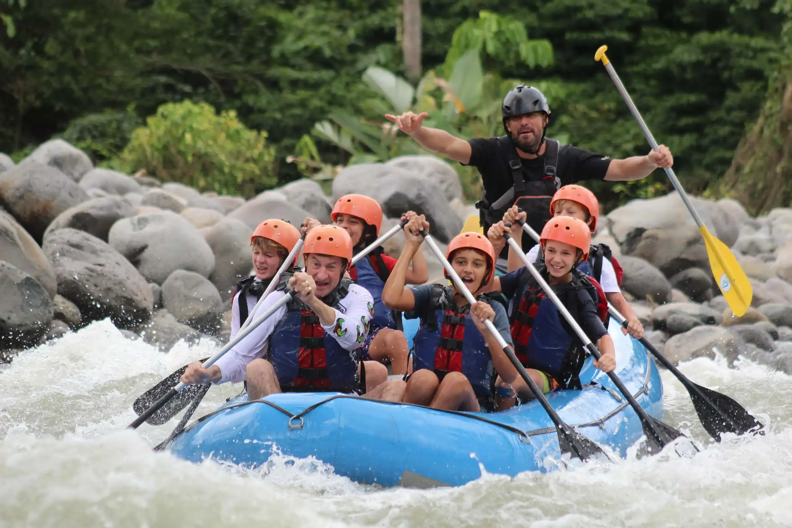 A group of people wearing helmets and life jackets paddle a blue inflatable raft through whitewater rapids, with a guide steering at the back—an exciting service travel for teens adventure.
