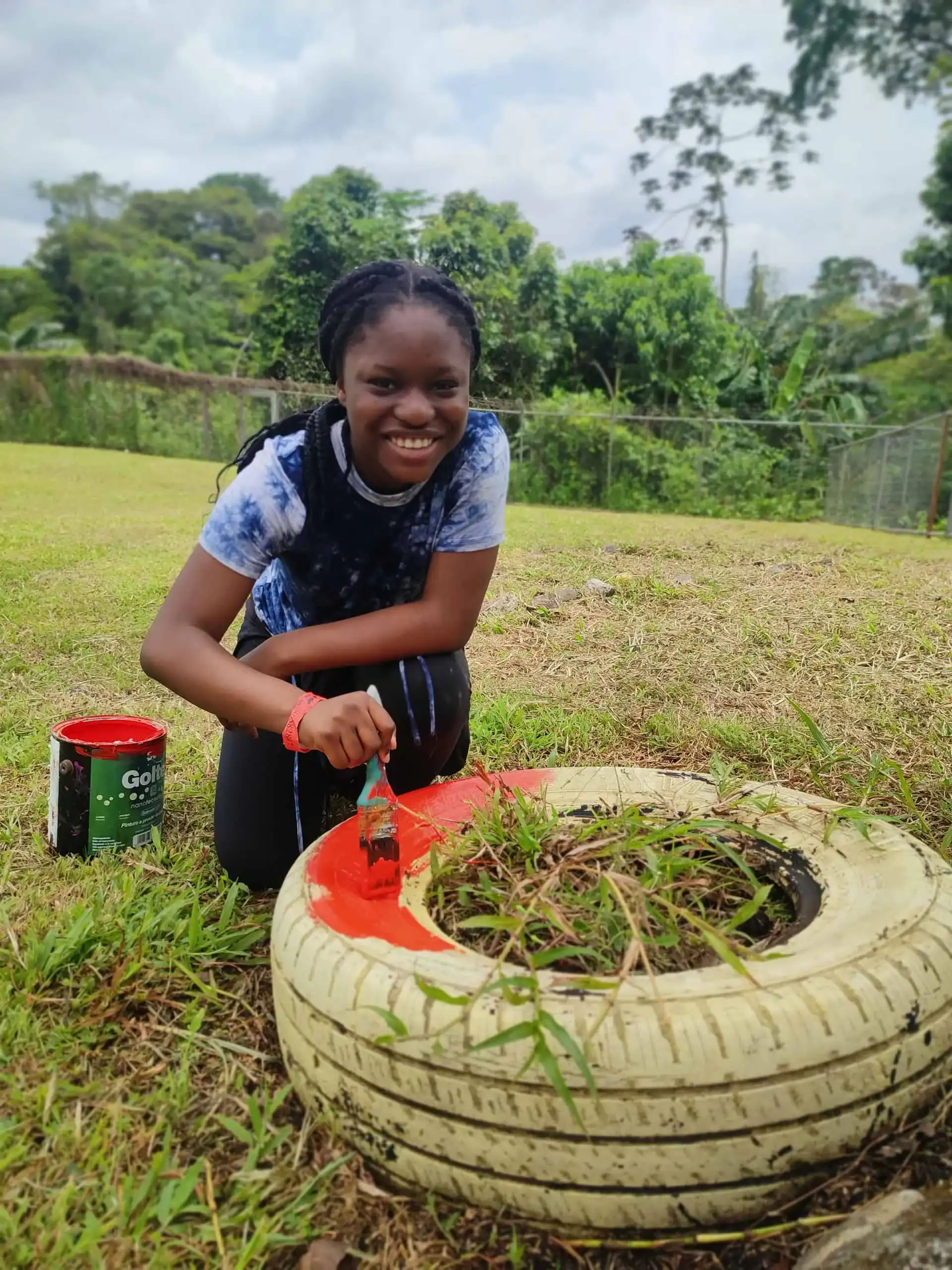 A person smiles while painting the edge of a white tire planter orange in an outdoor grassy area during a Costa Rica school trip, with a paint can nearby.
