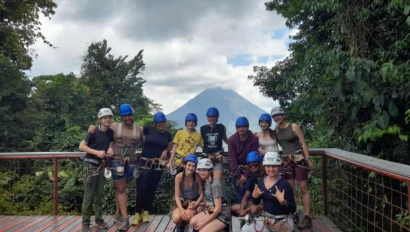 A group of people in helmets and harnesses pose on a wooden deck with a volcano and dense forest in the background.