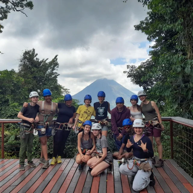 A group of people in helmets and harnesses pose on a wooden deck with a volcano and dense forest in the background.