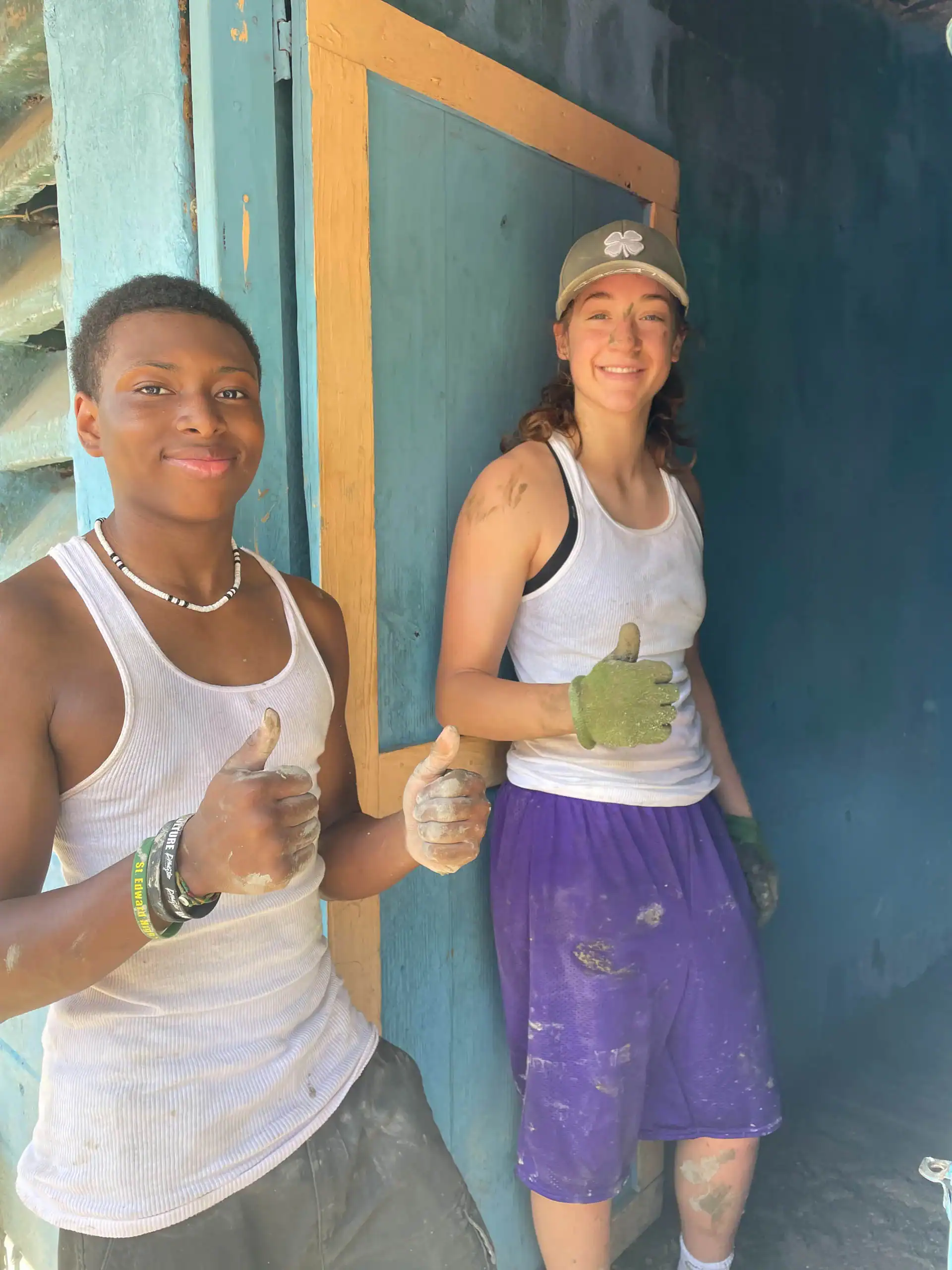 Two people wearing work clothes and giving thumbs up, standing by a blue wooden wall with paint and dirt on their clothes and hands after a successful Dominican Republic school trip project.