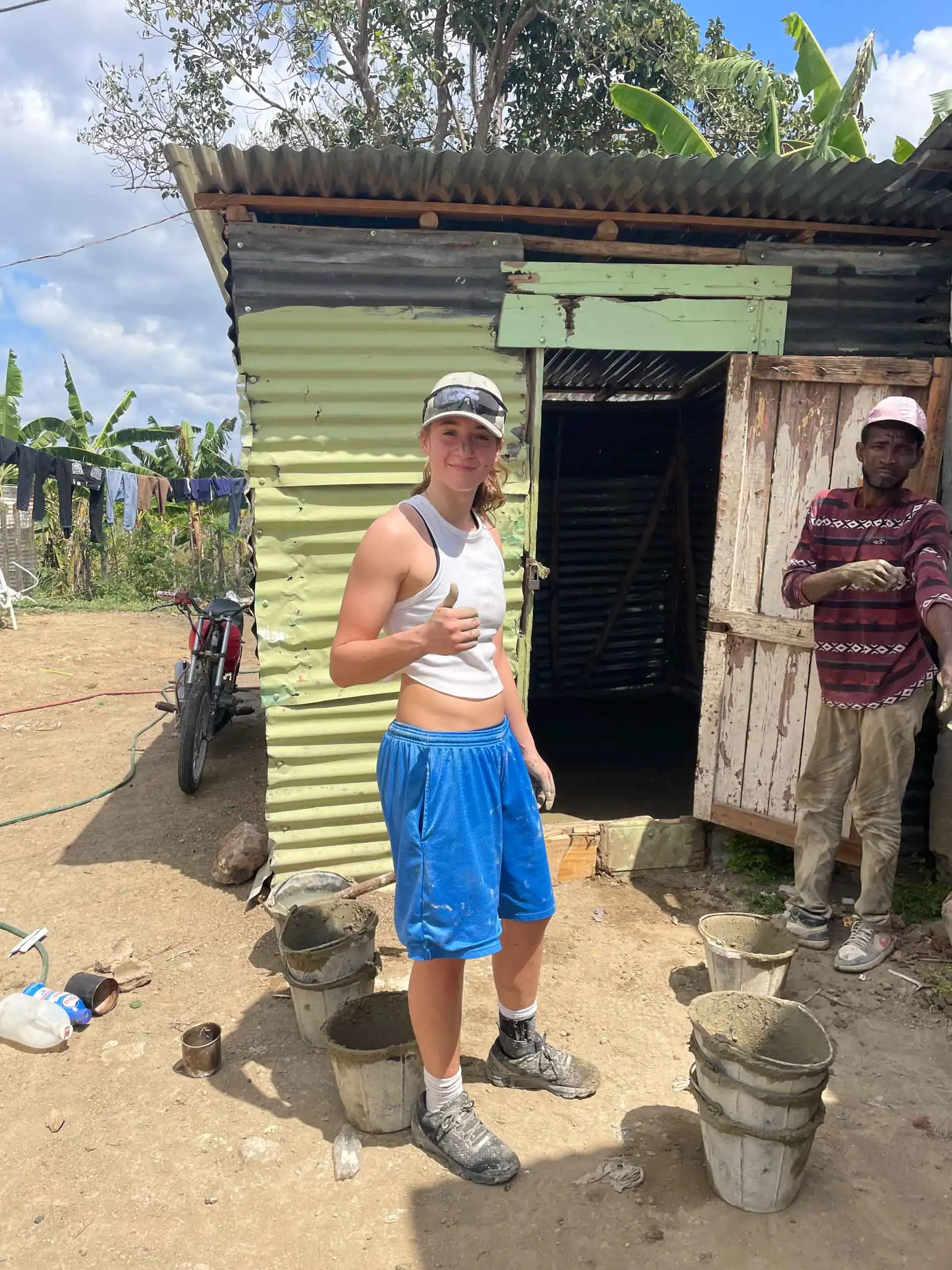 A person in a white tank top and blue shorts stands outside a corrugated metal building, giving a thumbs up during a service travel for teens trip, with several buckets on the ground and another person nearby.