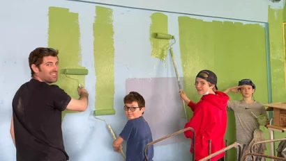 During Ecuador student travel, one adult and three children use paint rollers to paint a blue wall green inside a room with a corrugated metal roof.