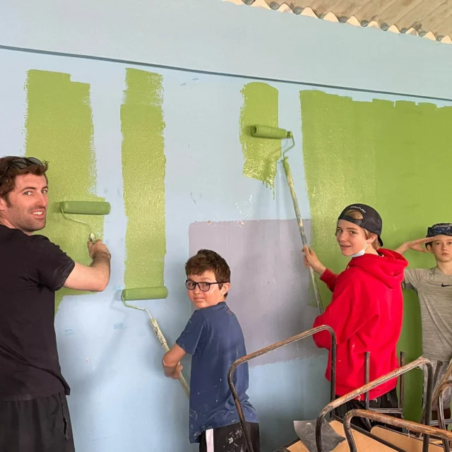 During Ecuador student travel, one adult and three children use paint rollers to paint a blue wall green inside a room with a corrugated metal roof.