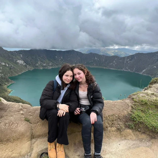 Two people sitting on a stone ledge with a scenic backdrop of a large blue lake surrounded by hills and cloudy skies.