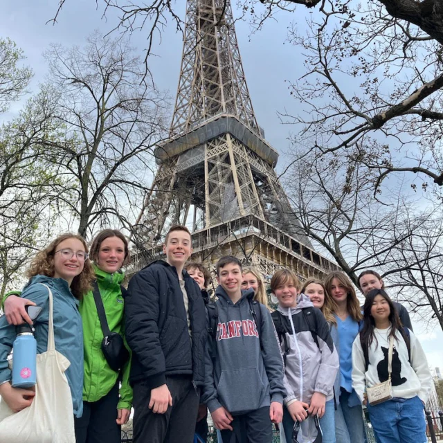 A group of people stands in front of the Eiffel Tower, with bare trees in the background. Some are holding bags and water bottles.