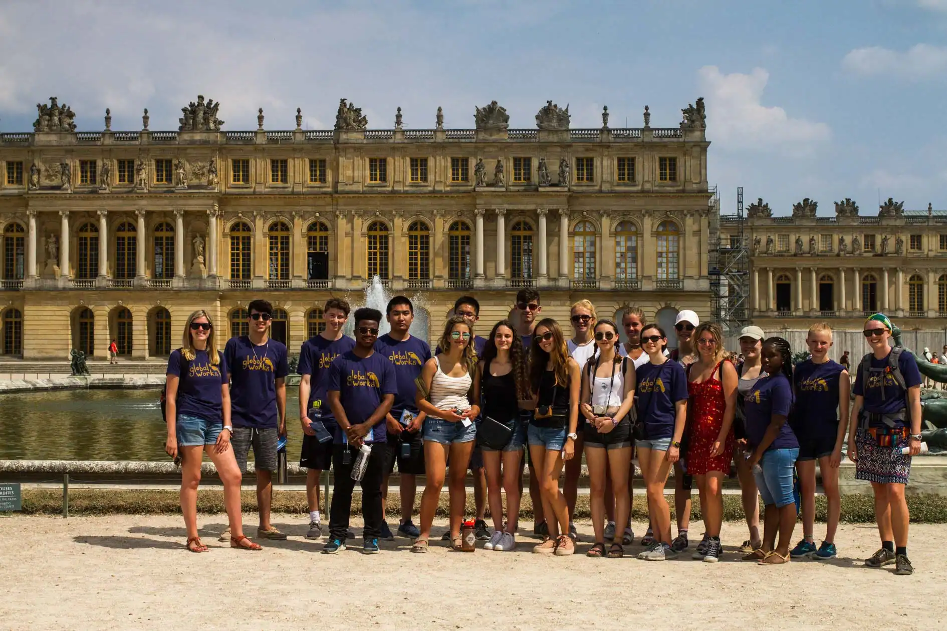 A group of young people on a France school trip pose in front of the Palace of Versailles, with some wearing matching purple shirts, on a sunny day.