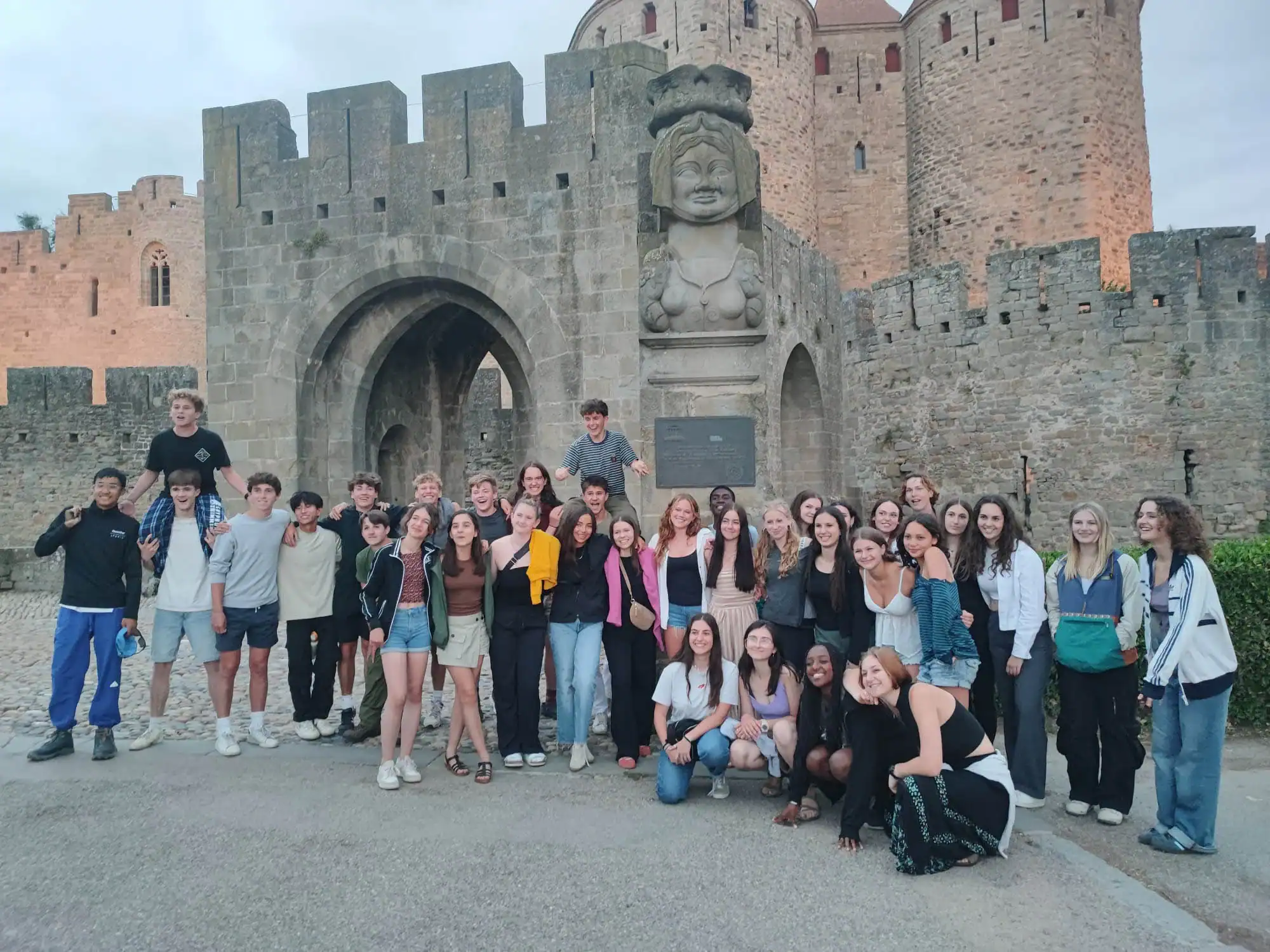 A group of young people on a France school trip pose for a photo in front of a stone castle entrance, featuring an arched doorway and a large stone bust.