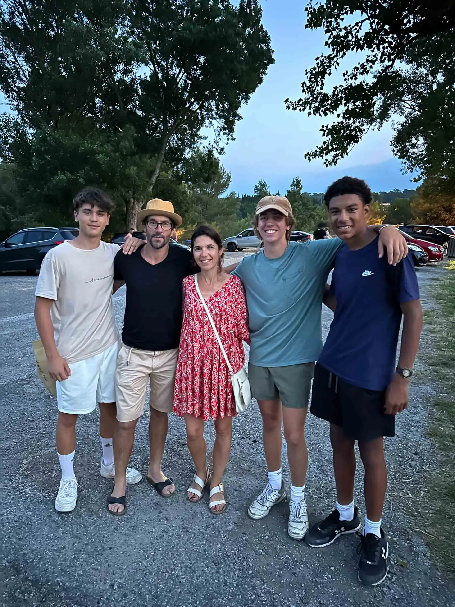 Five people stand together outdoors in a parking lot, posing for a group photo during their France school trip; trees and parked cars are visible in the background.