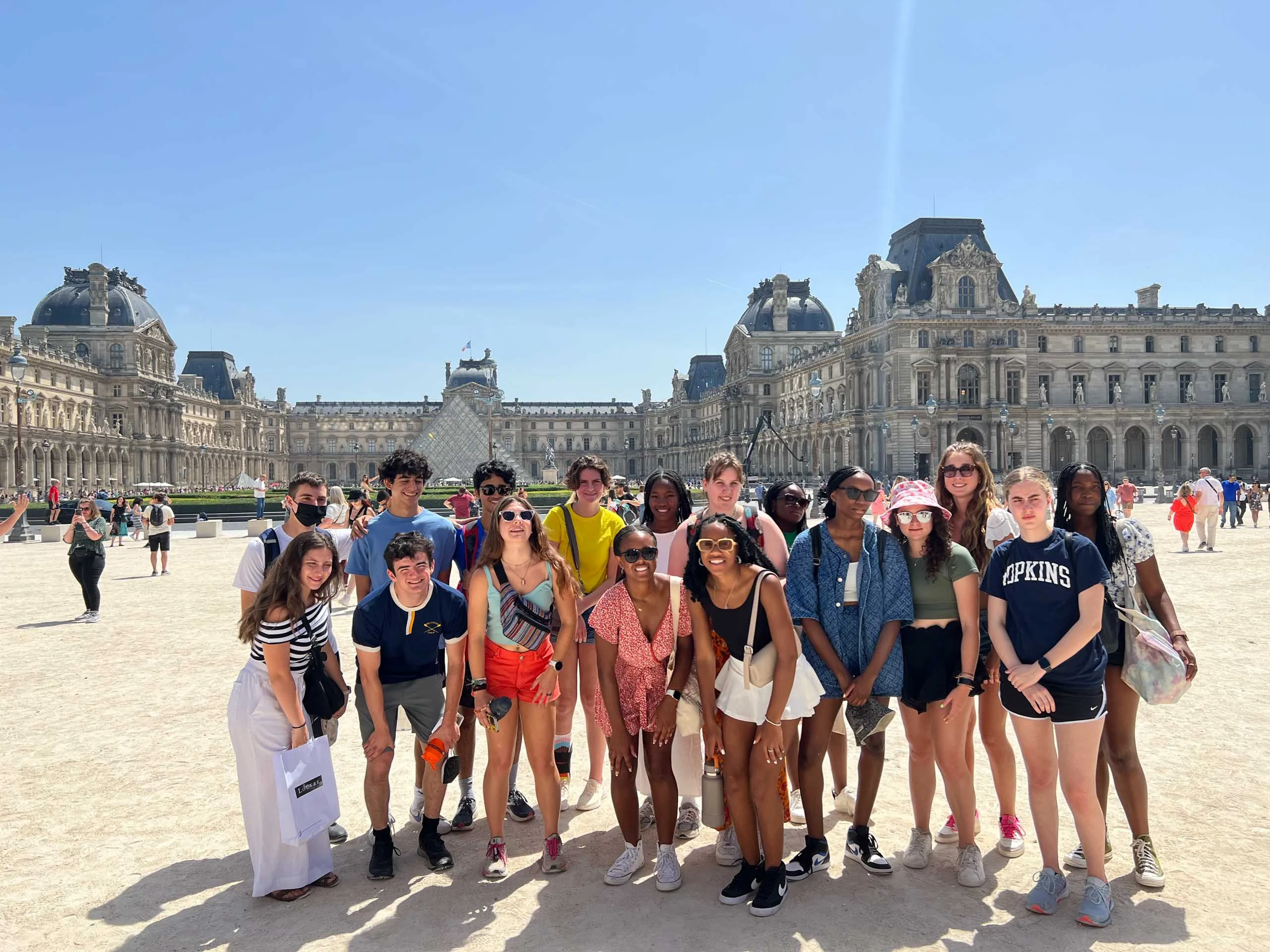 A group of young people poses for a photo outside the Louvre Museum in Paris on a sunny day, capturing memories from their France school trip with the historic building visible in the background.