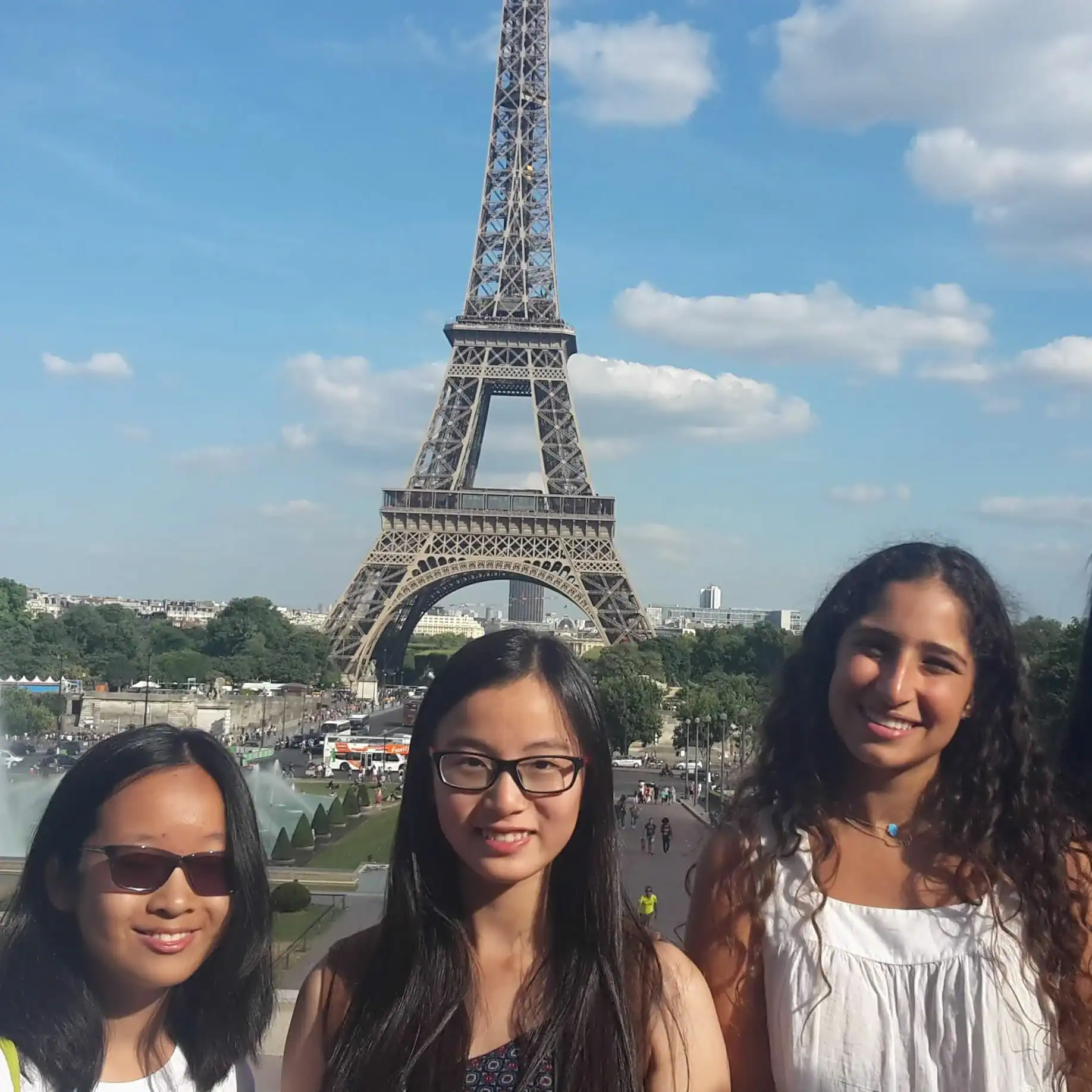 Three students stand in front of the Eiffel Tower in Paris on a sunny day with a blue sky and scattered clouds, capturing memories from their France school trip.