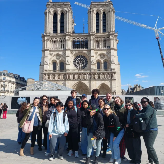 Group of people posing in front of the Notre-Dame Cathedral in Paris on a sunny day. Construction cranes are visible in the background.