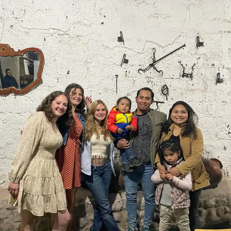 Six people, including two small children, pose for a photo during their language immersion trip, standing against a textured white wall decorated with rustic tools and a mirror.