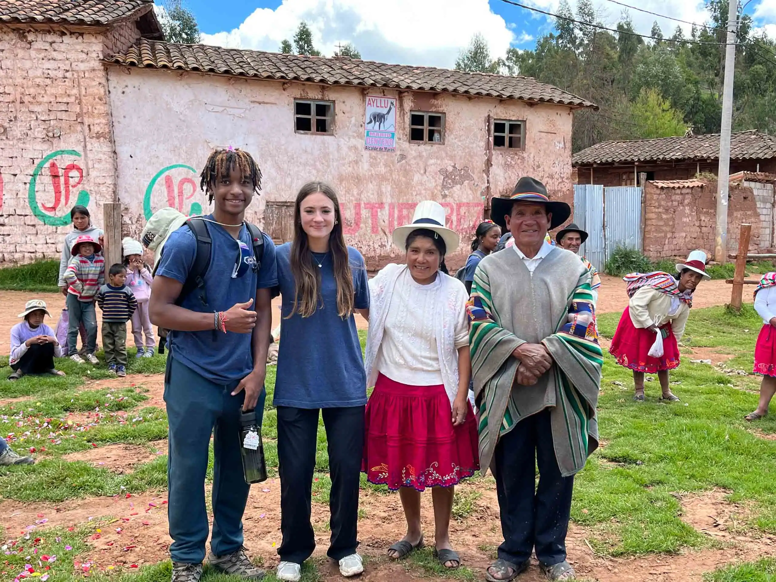 A group of four people stands together outside in front of a rustic building during a Peru school trip, with several others, including children, visible in the background.