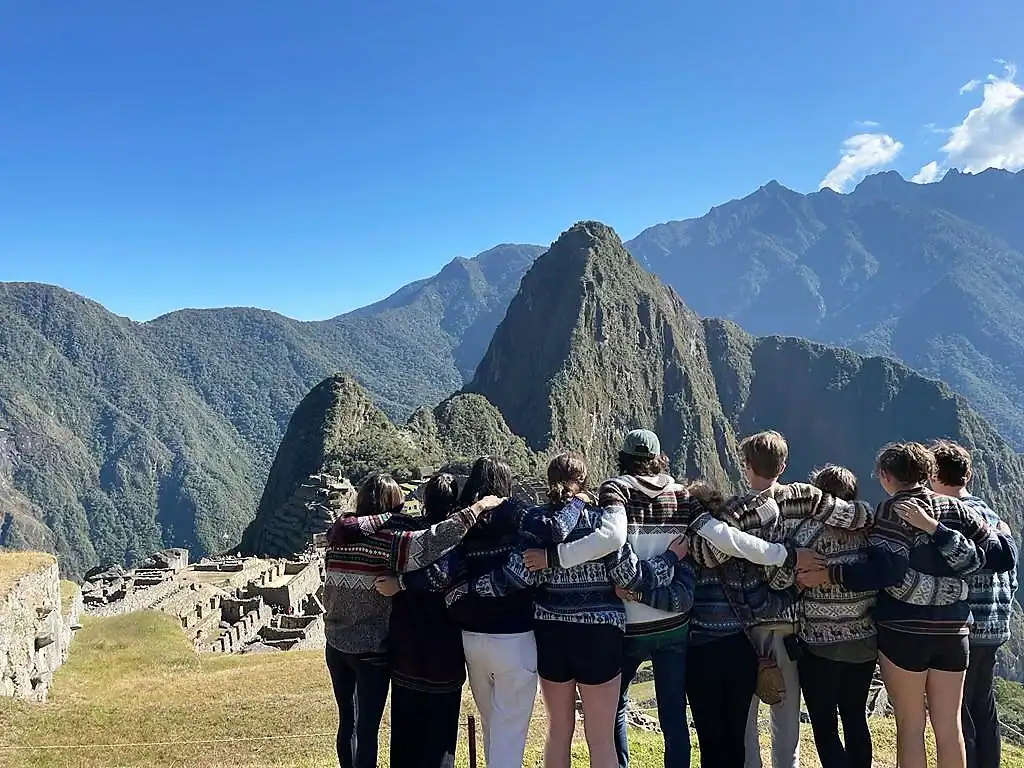 A group of students stand arm in arm facing Machu Picchu, with green mountains and a clear blue sky in the background, capturing the spirit of a memorable Peru school trip.